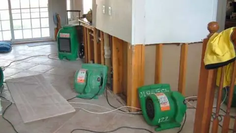 Green air movers and a dehumidifier drying a room where the lower wall drywall has been cut away